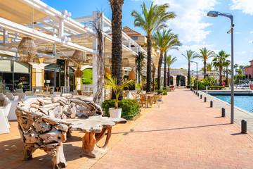 Restaurant tables with chairs in beautiful Sotogrande marina with colorful houses and palm trees on coastal promenade, Costa del Sol, Spain