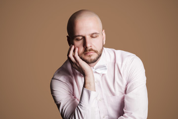 Portrait of thinking handsome bearded bald man in light pink shirt and white bow, sitting on chair touching his face with hand and closed eyes. indoor studio shot, isolated on brown background.