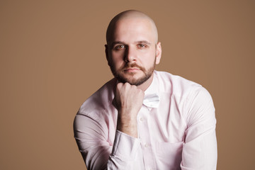 Obraz premium Portrait of thoughtful handsome bearded bald man in light pink shirt and white bow, sitting on chair and looking at camera with serious thinking face. indoor studio shot, isolated on brown background.