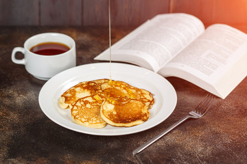 Pancakes with honey on white plate, tea and book