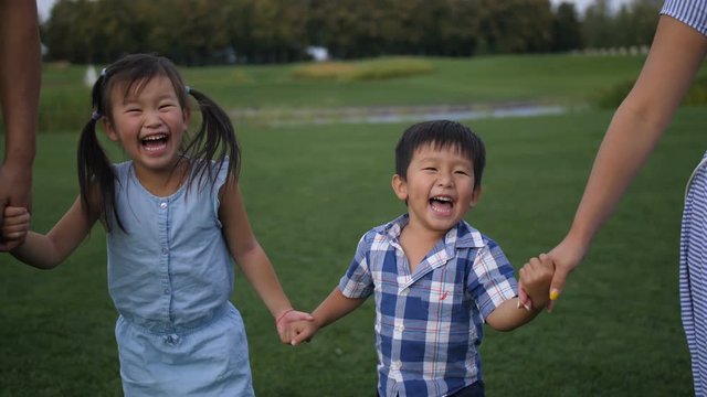 Adorable laughing asian siblings holding hands with parents and walking in summer park. Excited cute preschool girl with pigtails with her younger toddler brother enjoying a walk during family weekend