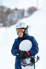 Fototapeta na wymiar cute young boy in gray helmet and orange googles, in blue jacket holds snowboard on white snow background. winter sport, active lifestyle, snowboarding concept 