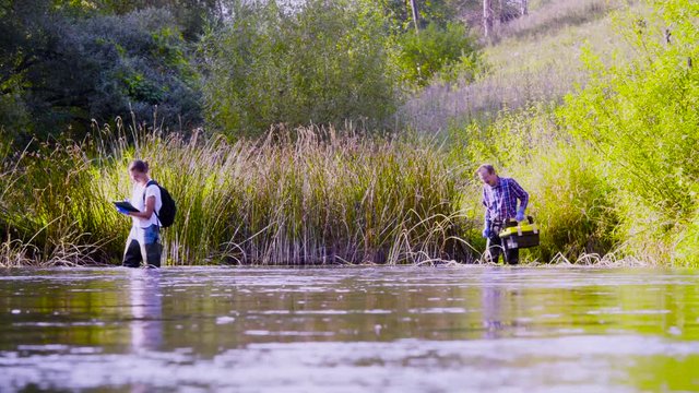 Crane shot. Two scientists ecologists in high rubber boots walking in the water of the forest river. Field work