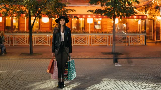 Time-lapse of young woman shopaholic standing outdoors in the street with shopping bags and looking at camera while flow of people is moving around her.
