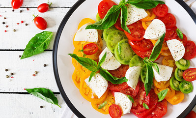Mozzarella cheese, tomatoes and basil herb leaves in plate on the white wooden table. Caprese salad. Italian food. Top view