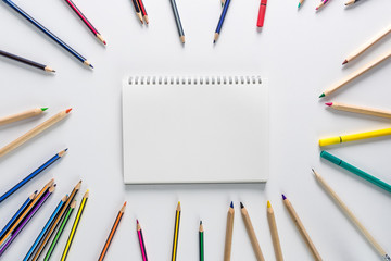 Top view over a school supplies as coloured pencils placed in a circle on a white background with blank work book in the middle of it. Back to school concept. Office supplies flat lay