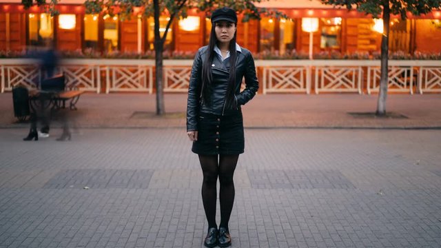Time-lapse portrait of serious Asian woman in stylish clothes standing in the street in the evening and looking at camera when crowds of men and women are passing by.