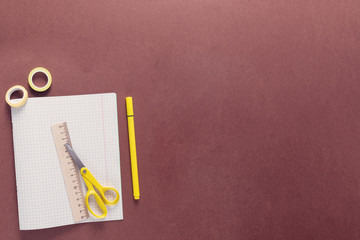 Top view over a school supplies as pens, work book, tape, scissors, ruler from left side of a brown background. Back to school concept. Office supplies flat lay