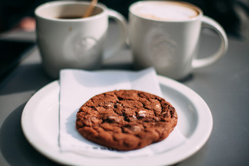 Cozy coffee break with chocolate cookie on white plate and two warm cups in soft café light, inviting morning treat close‑up