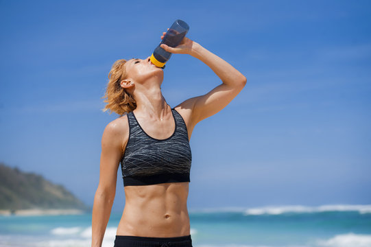 young happy and attractive sport runner woman drinking water bottle or isotonic drink after running workout at tropical paradise beach showing fit and athletic body