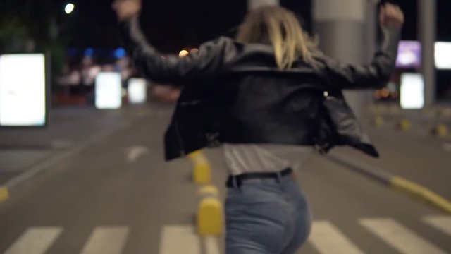 Young blonde woman in jeans and black leather jacket jumping on yellow street bumpers at night, having fun. Street, white city light, crosswalk on the background. Posing for camera, rare view