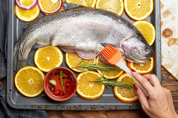 Fresh salmon lying on a baking sheet with oranges. Hand chef with a brush smears salmon oil. The concept of cooking salmon.