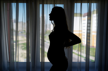 Fototapeta na wymiar Silhouette of a young pregnant woman who is standing in front of a window.