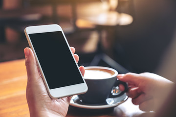 Mockup image of hands holding white mobile phone with blank screen while drinking coffee in modern cafe