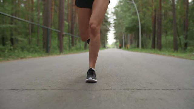Low-section dolly shot of legs of unrecognizable sportswoman running on pavement in park