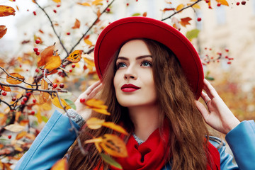 Close up outdoor portrait of young beautiful happy smiling girl wearing red hat and scarf posing near autumn tree. Model with red lips, long hair. Lady looking up.