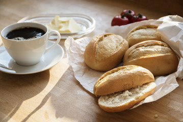 bread rolls or buns in a white paper bag, tomatoes, butter and a cup of coffee for breakfast on a...