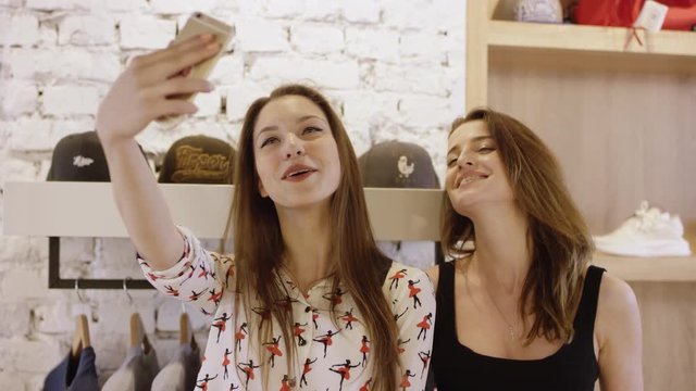 Portrait of young girls that are laughing using smartphone taking selfie in shopping mall. There are lights decoration and shelves on background.