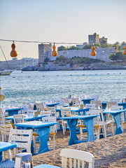 Obraz premium Tables with chairs of a Turkish tavern near the sea in a beach of Kumbahce bay with the Castle of Saint Peter in the background