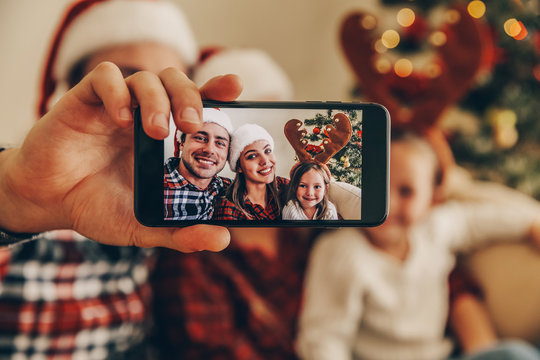 Christmas. Family. Technology. View on the smartphone. Dad, mom and daughter in Santa hats looking at camera and smiling while doing selfie