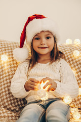 Christmas. Holiday. Portrait of little girl in Santa hat holding Christmas lights, looking at camera and smiling while sitting on couch at home