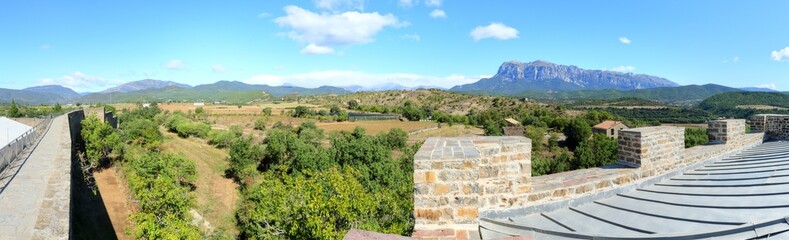 Fototapeta na wymiar A landscape with blue sky, some clouds, the medieval walls and the The Peña Montañesa mountain massif in the Spanish Aragonese Pyrenees, as seen from Ainsa, a small, stone made, rural town