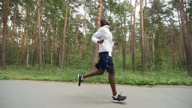 Tracking shot of tired black man in windbreaker and shorts jogging along forest road and laughing