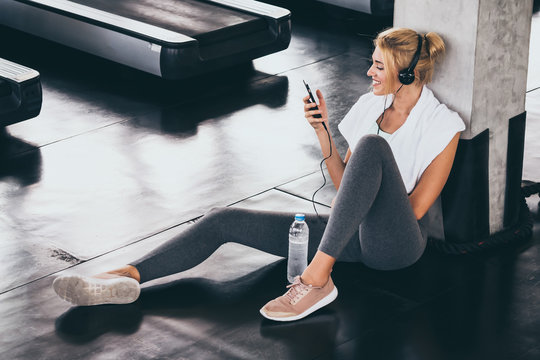 Young Happy Woman Sitting And Listening To Music By Smartphone While Having Exercise Break At The Gym.