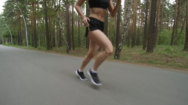 Tracking shot of young woman with muscular body running along road in park on gloomy morning