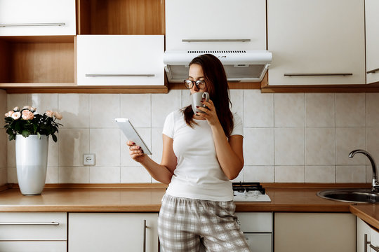 Home. Technologies. Relax. Girl in pajama and eyeglasses is using a digital tablet and drinking tea while standing in kitchen at home