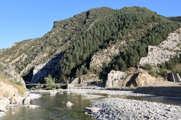 A landscape of Spanish Aragonese Pyrenees, with forest, mountains and an old wooden bridge which leads to Janovas, an abandoned town due to the construction of a dam. The river is Rio Ara.