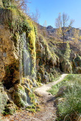 A small waterfall covered with moss next to a path in backlight during autumn in the Aguallueve rural park in Anento, Aragon, Spain