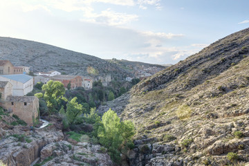A landscape at the sunset of the rural town of Almonacid de la Cuba, in Spain, with houses on a small valley with the Aguavivas river and waterfall. An ancient Roman bridge is crossing the river.