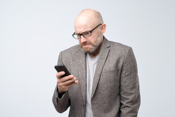 Dissatisfied man in casual suit wearing glasses reading articles, doing searching for information using phone.