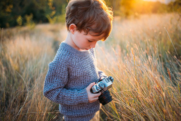 Cute toddler boy with old retro vintage camera on autumn grass background. Child with curly hair and grey coat playing with photo - Powered by Adobe
