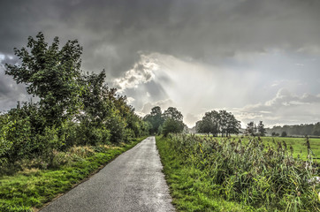 Narrow asphalt road between meadows and low forests near Ankeveen, The Netherlands with some raindrops falling from a diverse and rapidly changing sky