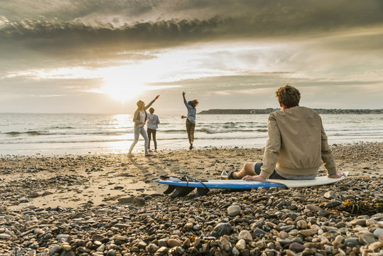 Young man on surfboard watching friends making soap bubbles on the beach at sunset