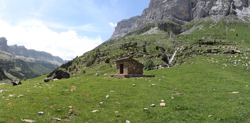 A landscape of small hut in the high mountains and massifs, pine and firs forest and a deep blue sky with some clouds in Ordesa valley (Valle de Ordesa) national park, in Aragon, Spain