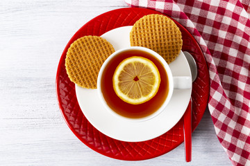 Tea in a white cup with lemon, Two waffles on a red plate. Napkin in a white and red cage. Tea spoon. White wood background. Top view. Copy space.