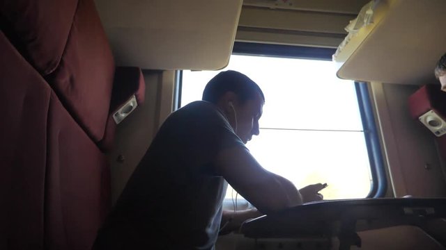 man silhouette travel is sitting on the train carriage holding sits by the window a smartphone Railway and drinking coffee and tea. slow motion video. man writes messages in the smartphone in the