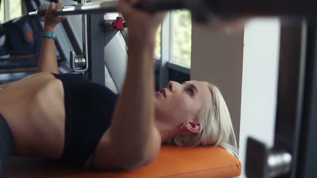 Close up footage of caucasian blonde woman in black sport bra training with bar on bench in the gym with panoramic windows. Concentrated on exercise, lifting clear bar. Side view