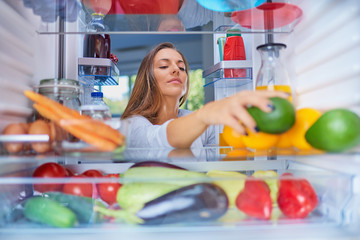 Woman standing in front of opened fridge and taking avocado. Fridge full of groceries.