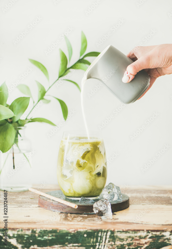 Poster iced matcha latte drink in glass with coconut milk pouring from pitcher by woman's hand, white wall 