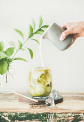 Iced matcha latte drink in glass with coconut milk pouring from pitcher by woman's hand, white wall...