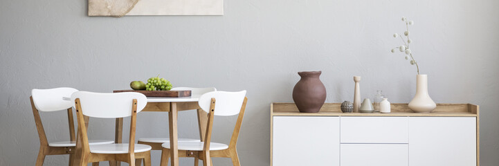Panorama of a cutting board with fruit on a simple white table with wooden chairs around in a bright dining room interior with gray walls