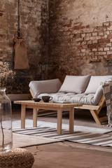 Wooden table in front of grey sofa in simple loft interior with rug and red brick wall. Real photo