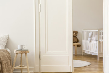 Real photo of wooden stool with book and tea mug standing next to white door to kid room interior with crib and herringbone parquet
