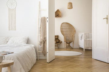 White apartment interior with herringbone parquet, double bed and open door to kid room with peacock chair, white crib and round rug on the floor