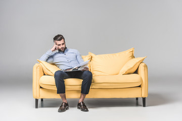 Fototapeta premium upset businessman reading newspaper while sitting on yellow sofa, on grey