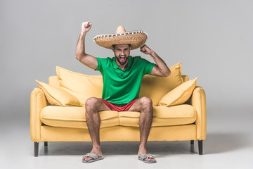 handsome man in mexican sombrero yelling and celebrating triumph on yellow sofa on grey